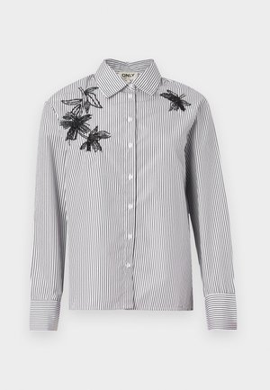 Camisa de botones a rayas en blanco y negro con mangas largas, que presenta bordados florales negros en el hombro izquierdo y la zona del pecho.