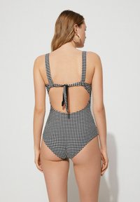 Černobílý kostkovaný jednodílný plavky s širokými ramínky, detaily na zavazování na zádech, výřezem na zádech a vysokými rozparky na nohou. Hladká textura.