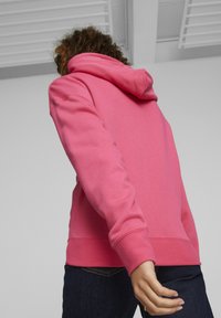 Sudadera rosa con capucha, de tejido suave, con puños y dobladillo acanalados. Llevada con jeans oscuros, mostrando la parte posterior y el brazo. Diseño simple, sin logotipos visibles.
