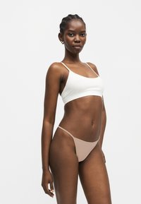 Γυναίκα με πλεξούδες που φοράει λευκό κοντό τοπ και μπεζ μαγιό κάτω μέρος, στέκεται μπροστά σε απλό λευκό φόντο.