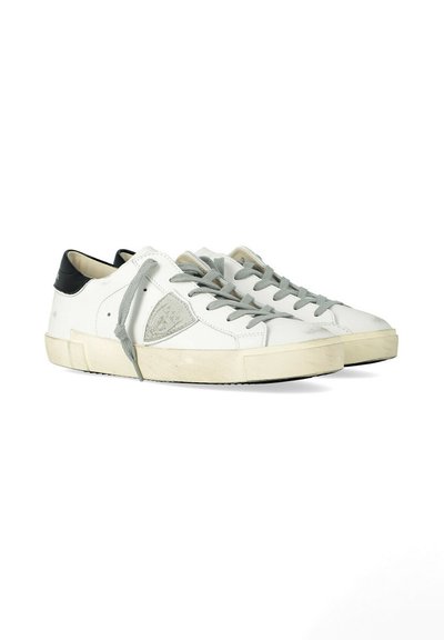 Baskets basses blanches avec lacets gris, semelles beige clair et languettes de talon noires, montrant une légère usure et un patch de logo cousu sur le côté.