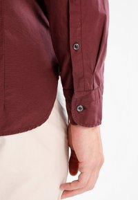 Detailní záběr na ruku člověka, která má na sobě dlouhý vínový rukáv s knoflíkovým manžetou a leží vedle světle béžových kalhot.