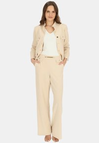 Beżowa marynarka dopasowana z złotymi guzikami, zestawiona z pasującym paskiem i szerokimi spodniami, noszona na białym topie z dekoltem w kształcie litery V. Gładka tekstura.
