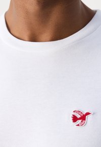 Weißes Baumwoll-T-Shirt mit Rundhalsausschnitt. Hat ein kleines, rotes, gesticktes Vogel-Logo auf der linken Brustseite. Glatte Textur.