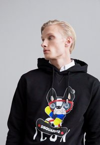 Svart huvtröja med en grafisk bild av en tecknad hund i skidutrustning. Har dragstrings och en ribbad textur, med "DSQUARED2 ICON" texten tydligt synlig.