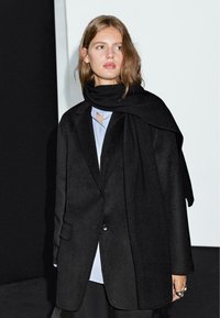 Černý oversized kabát s jedním knoflíkem, nošený přes světle modrou košili s výstřihem ve tvaru srdce. Měkce texturovaný šál přehnutý kolem krku.