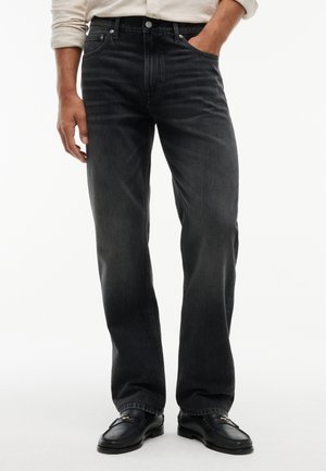 Uomo con jeans neri sbiaditi a gamba dritta, mocassini in pelle nera e una camicia chiara a maniche lunghe, una mano in tasca.