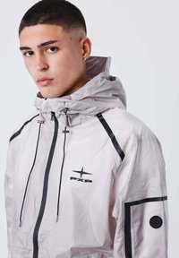 Giovane uomo con capelli corti che indossa una giacca a vento grigia chiara con zip nere e loghi PXP sul petto e sulle maniche.