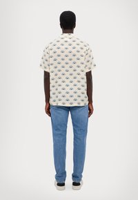 Korte mouw, crèmekleurig shirt met blauw oogpatroon, gecombineerd met lichtblauwe jeans en schoenen met zwarte accenten. De stof lijkt soepel en lichtgewicht.