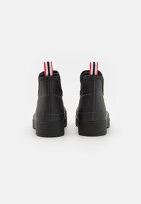 Bottes en caoutchouc noires à la cheville avec une finition lisse, dotées de poignées rayées rouges et grises, et d'une semelle plate épaisse.