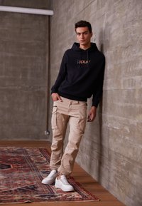 Sort hoodie med 'REFLX?' logo, beige cargo bukser og hvide sneakers. Modellen læner sig op ad en grå væg i en minimalistisk indstilling.