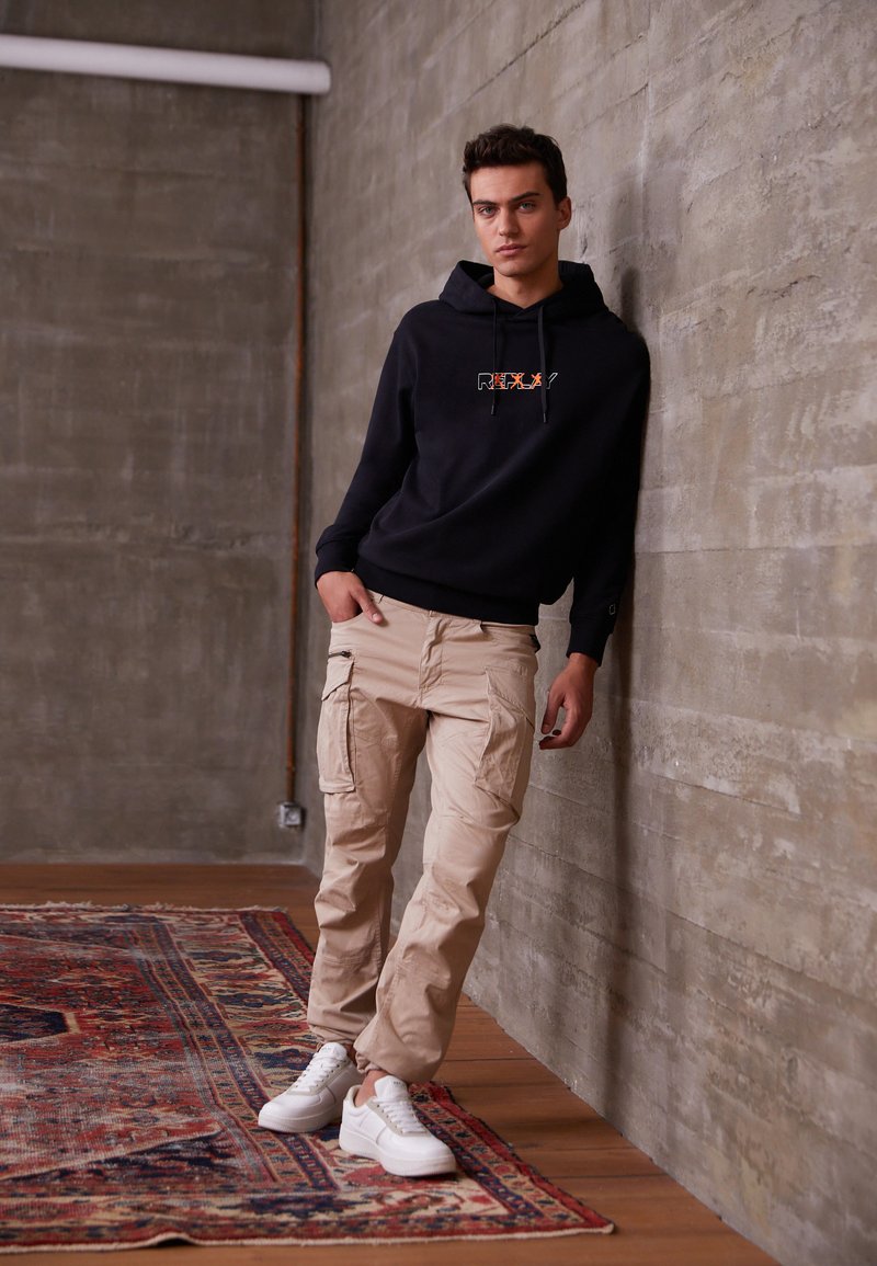 Sort hoodie med 'REFLX?' logo, beige cargo bukser og hvide sneakers. Modellen læner sig op ad en grå væg i en minimalistisk indstilling.