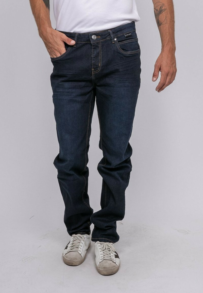 Jean en denim bleu foncé avec une coupe légèrement ample, des détails de couture visibles, un design à cinq poches et une fermeture à boutons. Porté avec des baskets blanches.