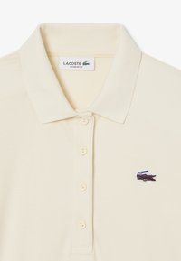 Polo en color crema claro, corte regular. Presenta un collar acanalado, tapeta de tres botones y un logo de cocodrilo bordado en color púrpura en el lado izquierdo del pecho.