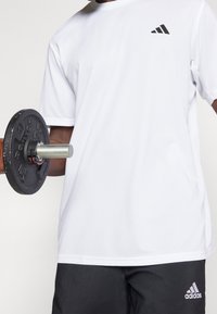 Camiseta atlética blanca de manga corta con un acabado texturizado y un logo negro de adidas en el lado izquierdo del pecho, combinada con pantalones cortos negros y un mancuerna.