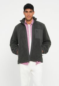 Jauns vīrietis valkā tumši pelēku flīsa jaku virs rozā kreklu ar pogām un baltām biksēm, stāvot pret baltu fona.