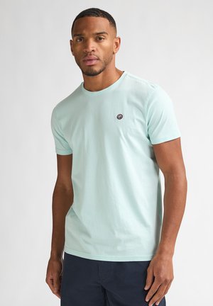 Mann trägt hellblaues kurzärmeliges T-Shirt mit kleinem Logo auf der Brust, steht vor einfachem hellgrau Hintergrund.
