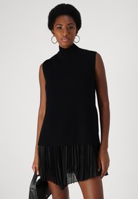 Μαύρο πλεκτό αμάνικο τοπ με ψηλό λαιμόκοψη, διαθέτοντας ριγωτή υφή, συνδυασμένο με μια πλισέ μαύρη φούστα. Το μοντέλο κρατάει μια μικρή μαύρη τσάντα.