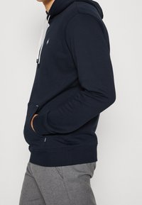 Sudadera con capucha de color azul marino hecha de tela suave, con un bolsillo frontal, puños acanalados y un parche con el logo en el pecho.