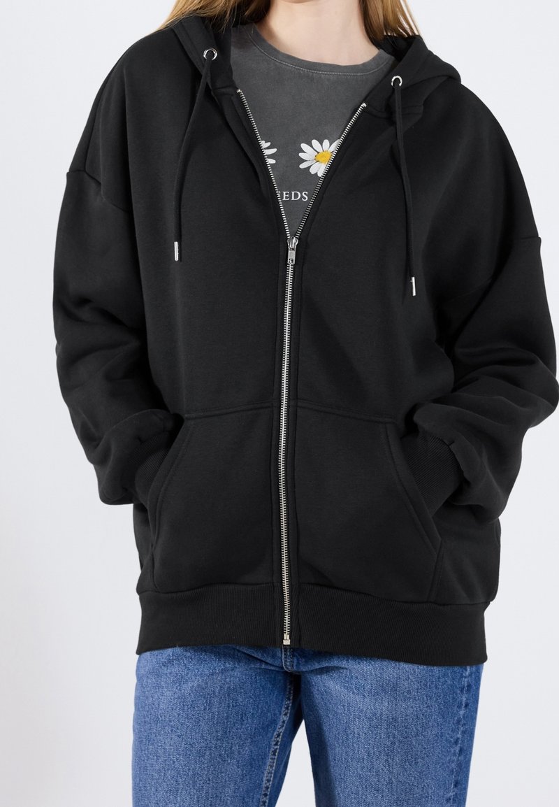Hoodie noir zippé avec cordons, grandes poches avant et poignets côtelés. Porté par-dessus un t-shirt gris à imprimé floral en dessous.