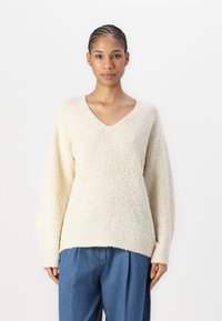Cream-colored, textured sweater with a V-neck and long sleeves. The fabric appears soft with a loose fit, paired with blue, pleated pants.