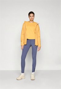 Aktyvaus poilsio apranga: geltona gobtuvuota striukė, geltona palaidinė, tamsiai mėlynos tamprės su siūlėmis ir šviesios sportinės batai.