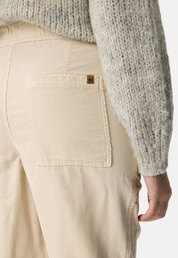 Pantalon en velours côtelé beige avec poche arrière et étiquette brune, associé à un pull en grosse maille gris clair vu de côté et de dos sur une personne.
