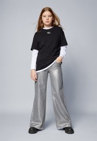 Tricou din bumbac negru peste o cămașă albă cu mâneci lungi; pantaloni lărgi din satin argintiu; pantofi masivi negri; detaliu simplu cu logo pe tricou.