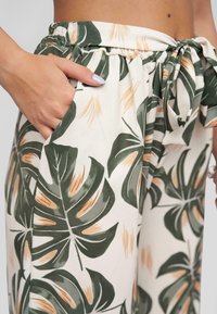 Pantalones blancos con estampado de hojas tropicales en verde y beige, que cuentan con una cinturilla elástica, un lazo decorativo y bolsillos laterales.