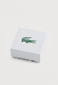 Balta kvadrātveida kaste ar teksturētu virsmu, uz kuras atrodas zaļas krāsas krokodila logotips un apakšā zaļā krāsā uzraksts "LACOSTE".