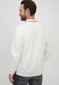 Langærmet polo skjorte i hvid bomuld med røde farver på kraven, der har en afslappet pasform og sideskår, parret med mørkegrå jeans.