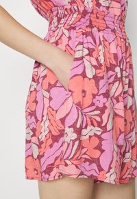 Main dans la poche d'une robe mi-longue à fleurs roses, violettes et oranges, taille froncée.