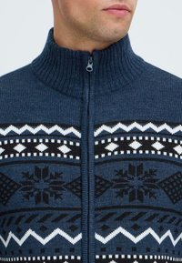 Jūrininkų megztinis su aukštu kaklu ir priekiniu užtrauktuku. Sukurtas su geometriniais raštais juodos ir baltos spalvų kombinacijoje krūtinės ir pečių srityje.