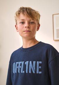 Puha pamutból készült tengerészkék pulóver, világoskék, texturált "OFFL1NE" felirattal. Klasszikus kerek nyakkivágás és bordás mandzsetták.