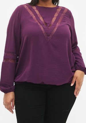 Kurvige Person trägt ein lila Langarmshirt mit transparenten Spitzendetails am V-Ausschnitt und an den Ärmeln, kombiniert mit einer schwarzen Hose.