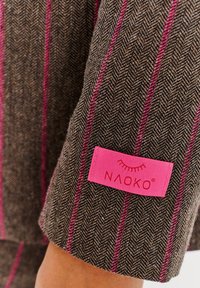 Látka s herringbone vzorem v hnědé barvě se růžovými pruhy; na manžetě je růžová visačka s nápisem „NAOKO“ červeným textem.