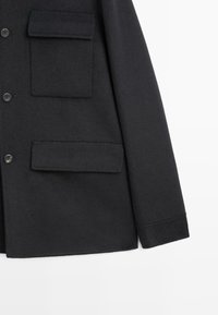 Cappotto in lana nera con finitura testurizzata, dotato di due tasche applicate frontali e chiusura a sei bottoni. Forma sartoriale con taglio pulito.