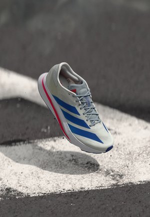 Scarpa da corsa grigio chiaro con strisce blu, dettagli rossi, tomaia strutturata e suola bianca, che sembra fluttuare sopra una superficie nera.