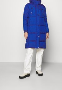 Blue puffer coat with a hood, featuring a quilted pattern, snap buttons, and two breast pockets. Paired with white pants and beige shoes.