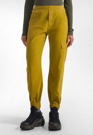 Person iført sennepsgule cargobukser med elastiske manchetter og sorte chunky sneakers stående mod hvid baggrund.