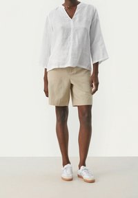 Personne portant un haut blanc à manches longues ample, un short beige jusqu'aux genoux et des baskets blanches avec des semelles en gomme, se tenant devant un fond uni.