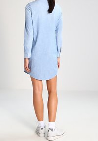 Světle modré šaty košilového střihu s dlouhým rukávem a zaobleným lemem, vyrobené z tkané látky; mají knoflíkové manžety a jsou kombinovány s bílými teniskami.