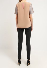 Žena v béžovém topu s krátkými metalickými rukávy a růžovým zipem na zadní straně, černých skinny kalhotách a černých vysokých podpatcích, stojí zády k nám.