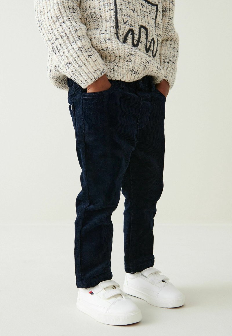 Enfant portant un pantalon en velours côtelé bleu marine, des baskets blanches à scratch, et un pull en tricot beige avec un motif graphique noir, les mains dans les poches.