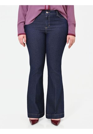 Donna che indossa jeans svasati blu scuro, una camicetta viola a maniche lunghe e scarpe rosse con tacco a punta, in piedi su sfondo bianco.