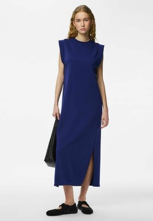 Noor naine, kellel on pikad lokkis juuksed, kannab varrukateta tumelilla midi-pikka kleiti õla patjadega, musti kingi ja hoiab suurt musta kotti.