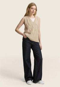 Gilet senza maniche in maglia beige con motivo a trecce, design con scollo a V, abbinato a jeans a gamba larga in denim scuro e sneakers bianche.