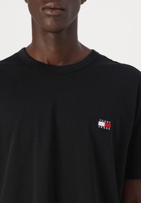 Close-up af en person iført en sort T-shirt med et lille rektangulært Tommy Jeans-logo på venstre bryst.