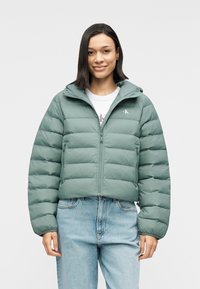 Πράσινο μπουφάν puffer με φερμουάρ, κουκούλα και λογότυπο στο στήθος. Διαθέτει σχέδιο καπιτονέ και ελαστικά μανίκια. Φοριέται πάνω από ένα λευκό μπλουζάκι και τζιν.