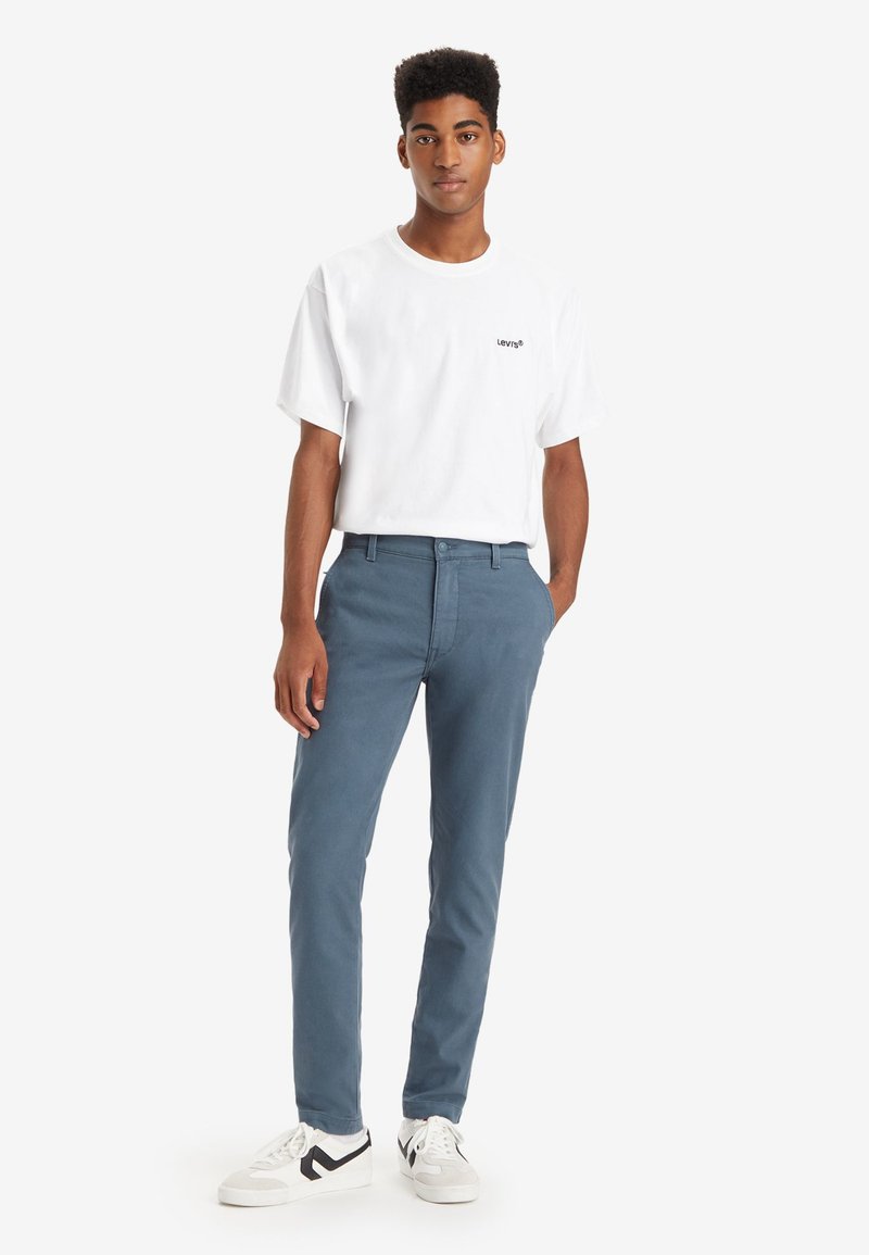 Jeune homme debout les mains dans les poches, portant un T-shirt blanc Levi's, un pantalon bleu et des baskets blanches avec des détails noirs, sur fond blanc.